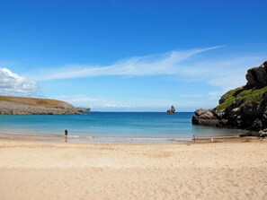 A beach with clear blue water, rocky cliffs, and a person walking on the sand.