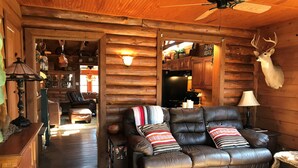 A cozy cabin interior with a leather sofa, wooden walls, a mounted deer head, and a kitchen area visible through an open doorway.