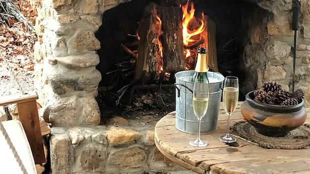 A stone fireplace with a fire, a wooden mantel, and a table with glasses and a bottle.