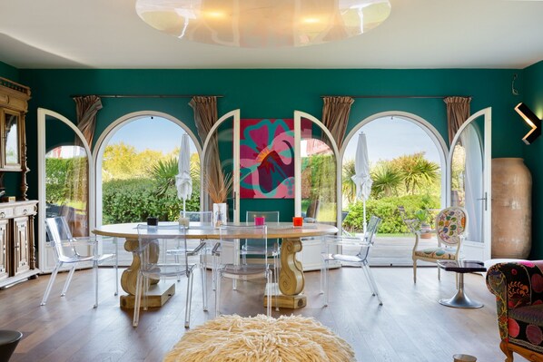 A dining area with a round table, clear chairs, and a view through large windows.