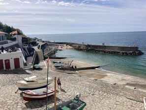 Eine Küstenlandschaft mit vor Anker liegenden Booten, einem mit Kopfsteinpflaster belegten Strand und einer Pier mit Personen.