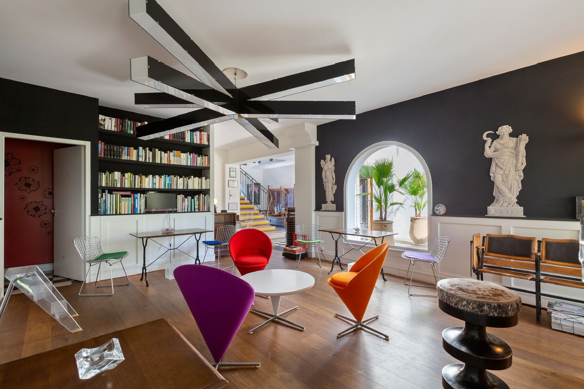 A modern living room with a bookshelf, colorful chairs, a round table, and a unique ceiling design.