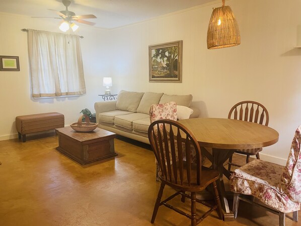 A living room with a sofa, a wooden coffee table, a dining table with chairs, a ceiling fan, and a hanging light fixture.