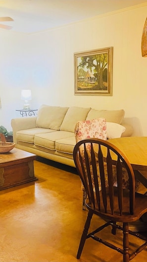 Una sala de estar con un sofá color crema, una mesa de comedor de madera y una silla de madera. Un cuadro de una casa bajo un árbol cuelga en la pared.