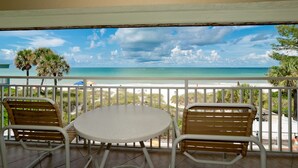 A balcony with two wooden chairs and a round table, overlooking a beach with palm trees and a clear sky. - Amazing View, Updated Beachfront Condo (Bradenton Beach)