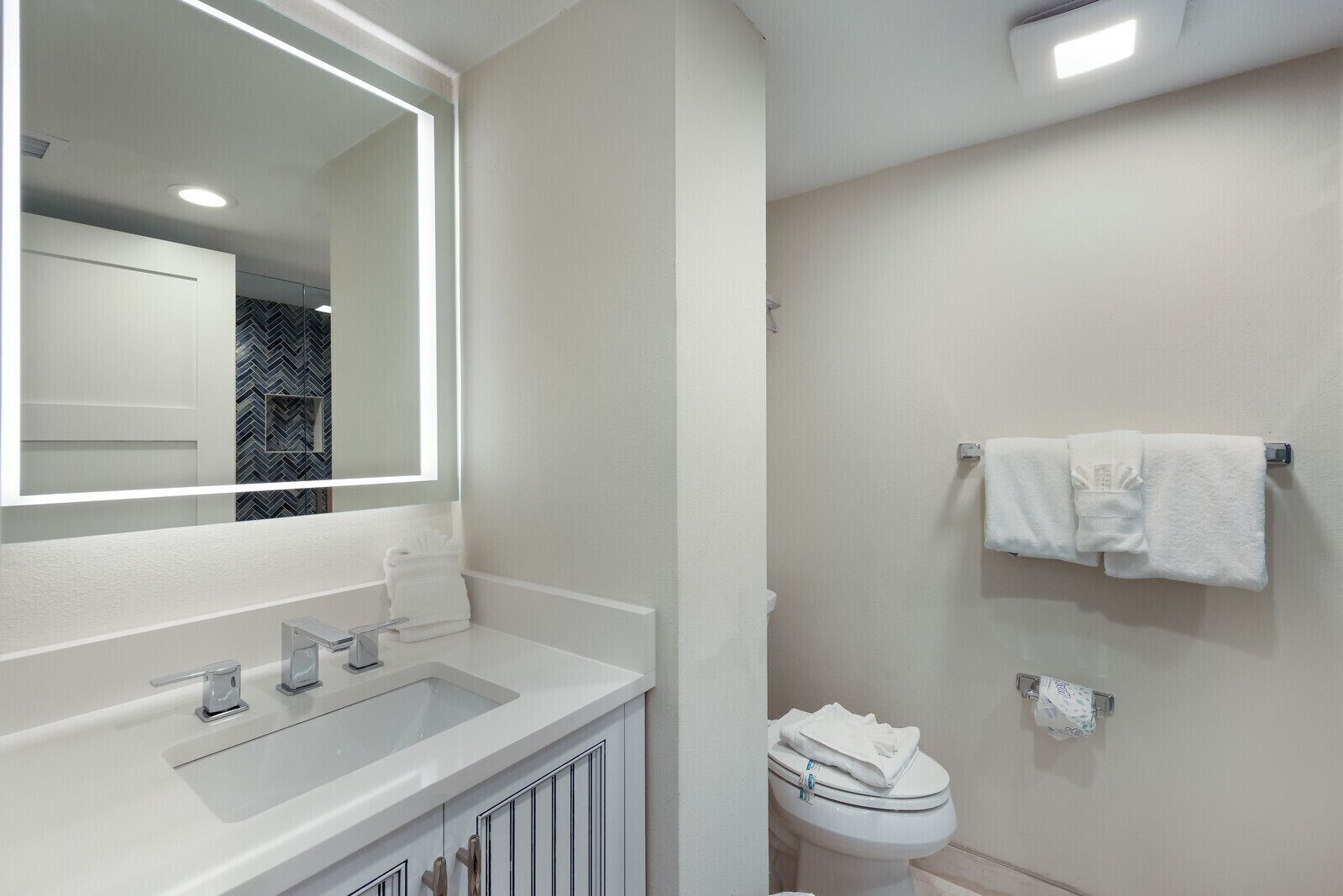 A modern bathroom with a white sink, mirror, and toilet. There are two towels on the wall and a toilet paper holder.