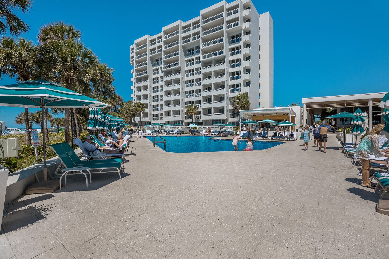 A resort pool area with sun loungers, umbrellas, and a large building in the background.
