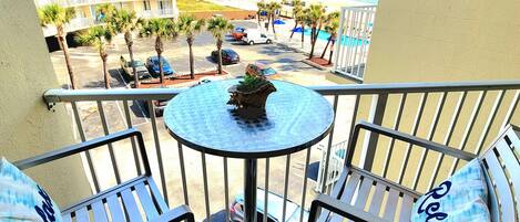 A balcony with a view of the beach, a pool, and a parking area with palm trees.