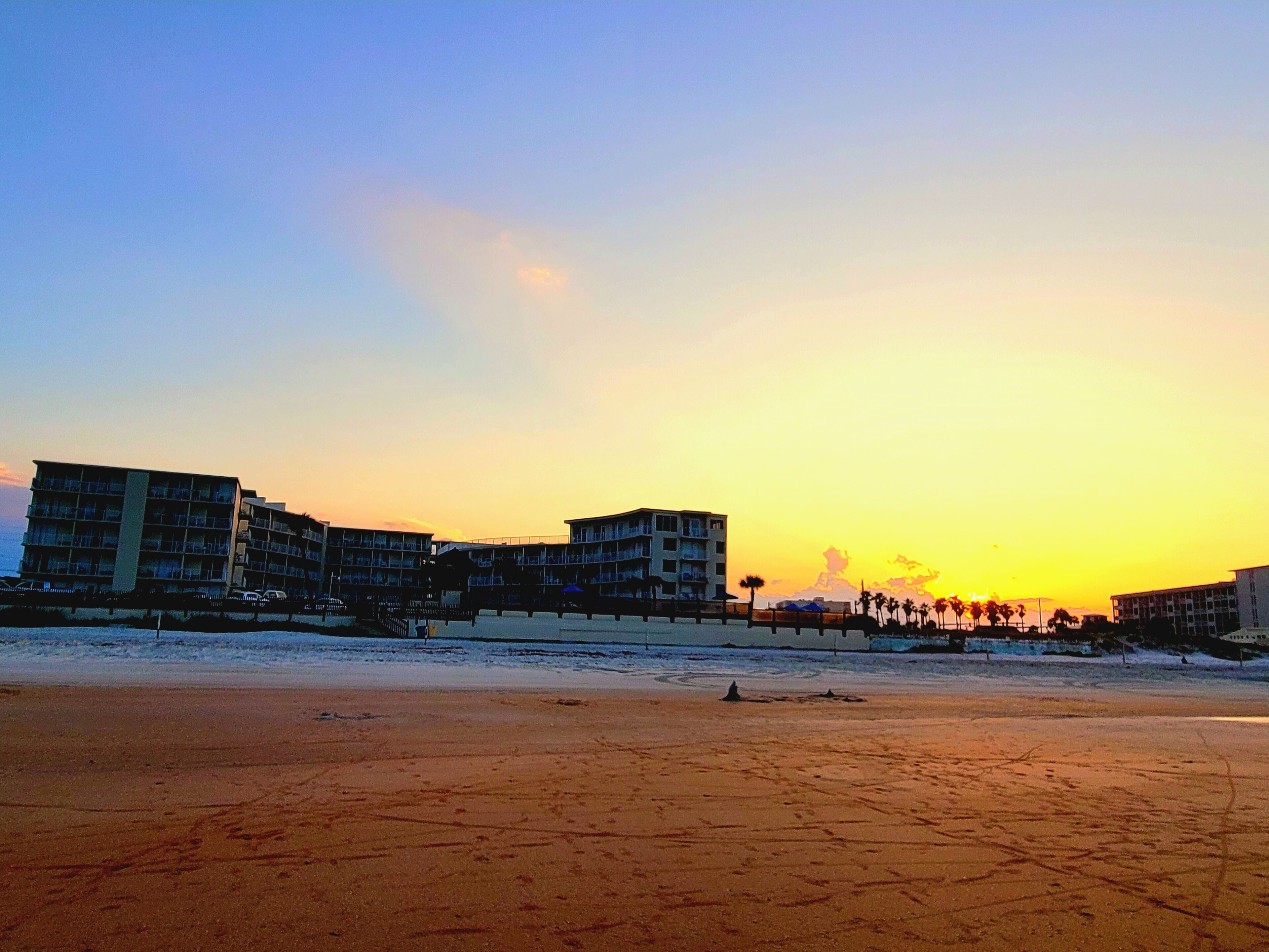 Ein Strandhotel mit mehreren Etagen und Balkonen, ein sandiger Strand im Vordergrund und Palmen im Hintergrund.
