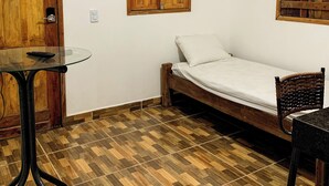 Et lille rum med en trædør, et glasbord med en fjernbetjening, en seng med hvid madras, en fletstol og en trægulv.
