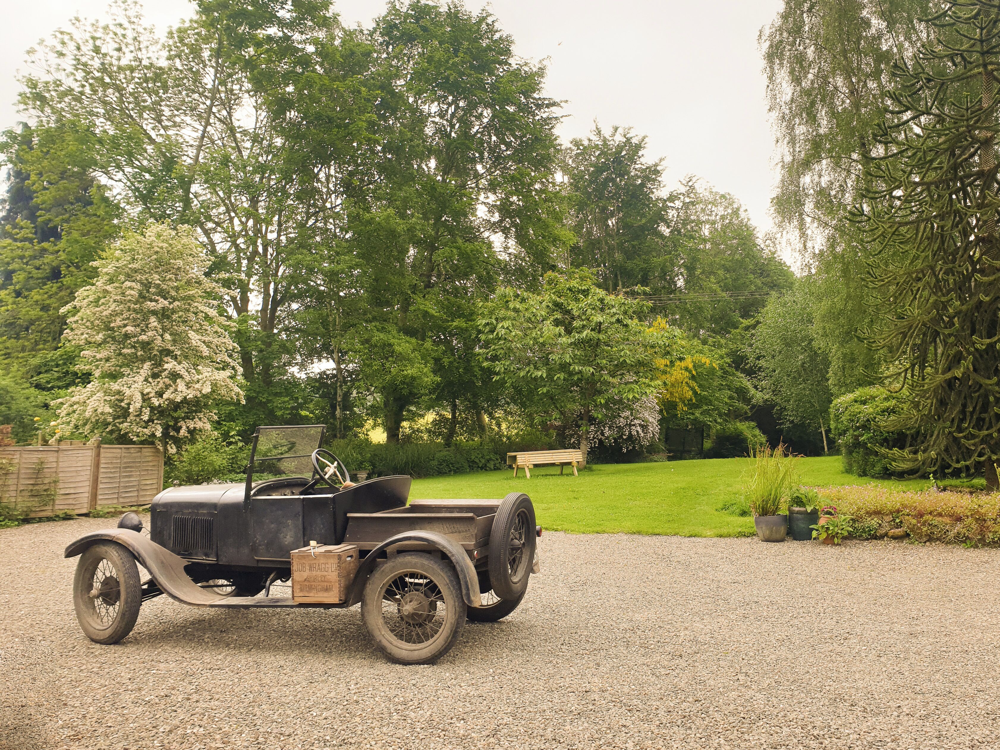 A vintage car parked on a gravel driveway in a lush garden with trees and a bench.