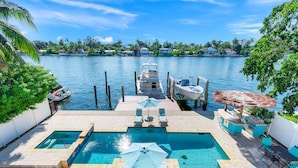 A swimming pool with lounge chairs and umbrellas, a dock with a boat, and a view of a water body with buildings in the background. - VIP Luxury 5 BR OceanFront Villa | Heated Pool & Dock | Free Parking & WiFi (North Bay Village)