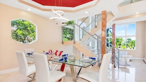 A modern dining area with a glass table, white chairs, and a staircase with glass railings. - VIP Luxury 5 BR OceanFront Villa | Heated Pool & Dock | Free Parking & WiFi (North Bay Village)
