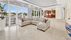 A spacious living area with a large sectional sofa, a dining area with a kitchen in the background, and a view of a marina through large windows. - VIP Luxury 5 BR OceanFront Villa | Heated Pool & Dock | Free Parking & WiFi (North Bay Village)