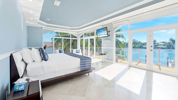 A modern bedroom with a large bed, a TV, and a balcony with a view of the water. - VIP Luxury 5 BR OceanFront Villa | Heated Pool & Dock | Free Parking & WiFi (North Bay Village)