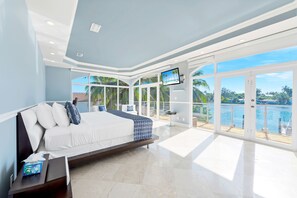 A modern bedroom with a large bed, a TV, and a balcony with a view of the water. - VIP Luxury 5 BR OceanFront Villa | Heated Pool & Dock | Free Parking & WiFi (North Bay Village)