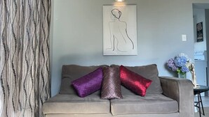 Ein graues Sofa mit drei Kissen in Lila, Braun und Rot. Darüber an der Wand eine Wandleuchte und ein gerahmtes Linienezeichnung.