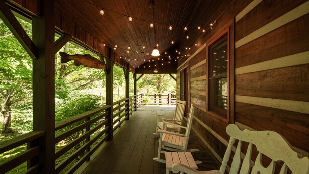 A covered porch with wooden railings, white wooden benches, and a view of a forested area. - Private, Peaceful Log Cabin w/ wrap around porch and fireplace. Central location (Boone)
