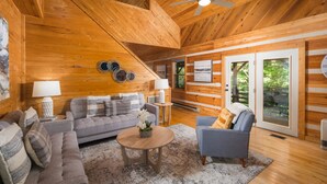 A cozy cabin interior with a wooden ceiling, a sofa, a chair, a coffee table, and a large window with a view of greenery. - Private, Peaceful Log Cabin w/ wrap around porch and fireplace. Central location (Boone)