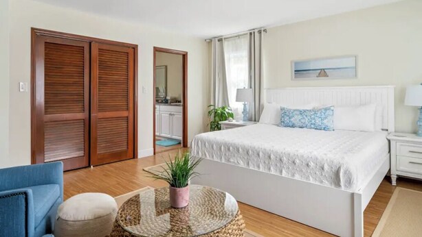 A bedroom with a large bed, a blue armchair, a round coffee table with a plant, wooden wardrobes, a nightstand, and a window with curtains. - Sea Shell Room — Bright Coastal Luxury with Extra Space to Unwind (Rehoboth Beach)
