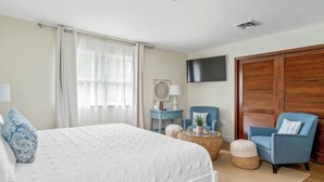 A bedroom with a bed, a TV, a chair, a side table, and a wooden wardrobe. - Sea Shell Room — Bright Coastal Luxury with Extra Space to Unwind (Rehoboth Beach)