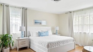 A bedroom with a bed, bedside tables, a nightstand, a potted plant, and a window with curtains. - Sea Shell Room — Bright Coastal Luxury with Extra Space to Unwind (Rehoboth Beach)