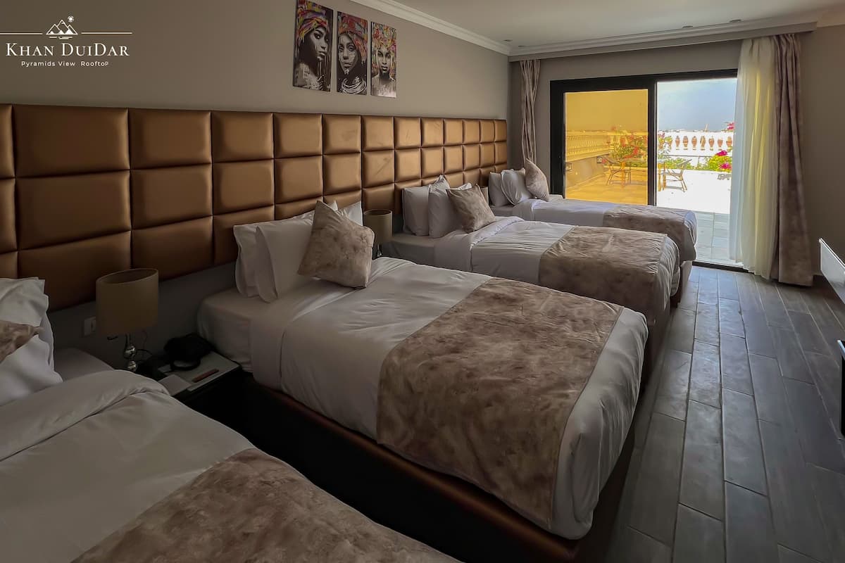 A hotel room with a large padded headboard, two beds with matching bedding, a bedside table with a lamp, and a view of the outdoors through a sliding door.