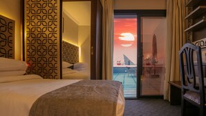 Habitació d'hotel amb llit, paret amb dibuixos, porta corredissa a un balcó i vista cap a un paisatge aquàtic al capvespre.