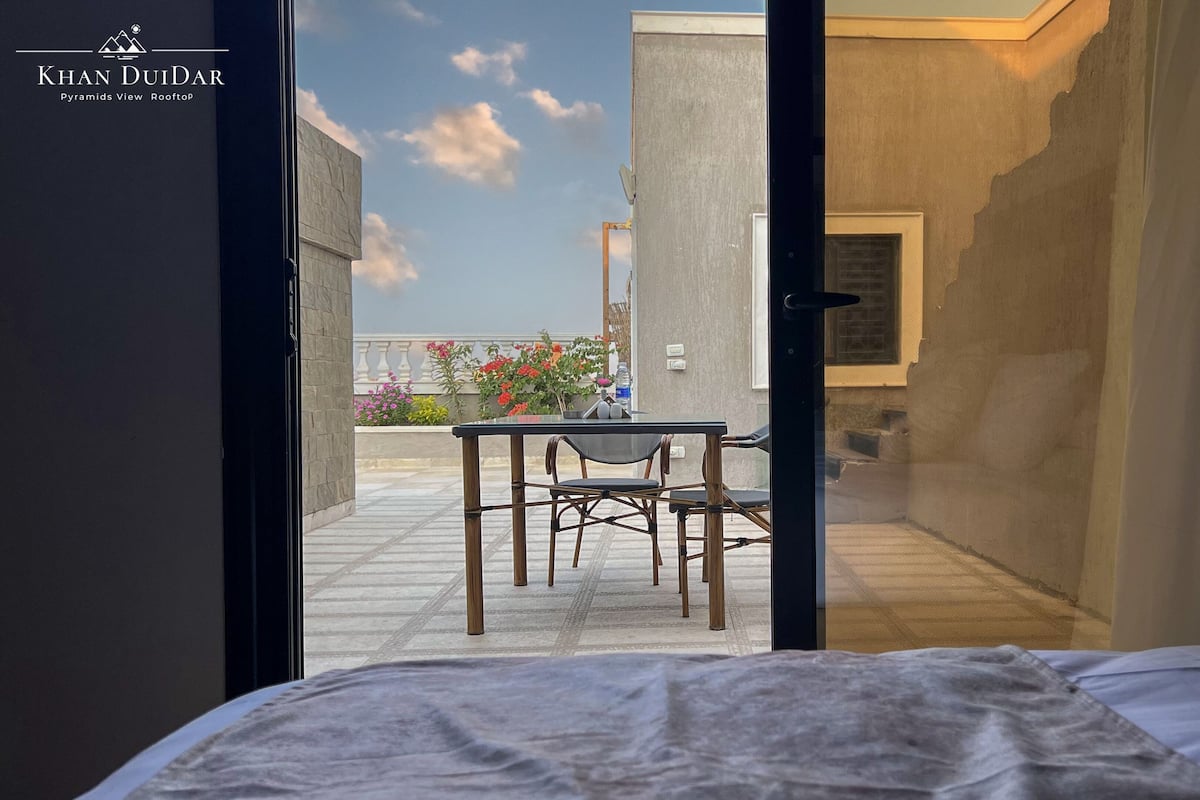 A hotel room with a bed, a table with chairs, and a view of the sky and flowers outside.