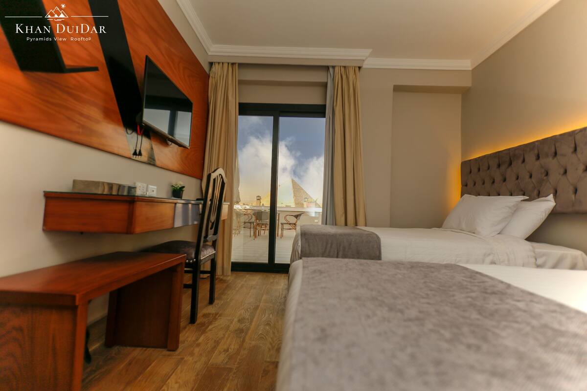 A hotel room with two beds, a desk, a chair, a TV mounted on the wall, and a view of the sky through the window.