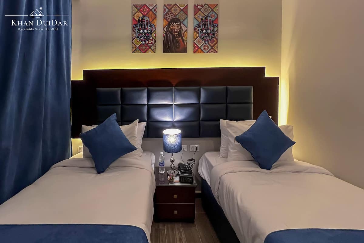 A hotel room with two beds, a nightstand, a lamp, and a view of the pyramids.