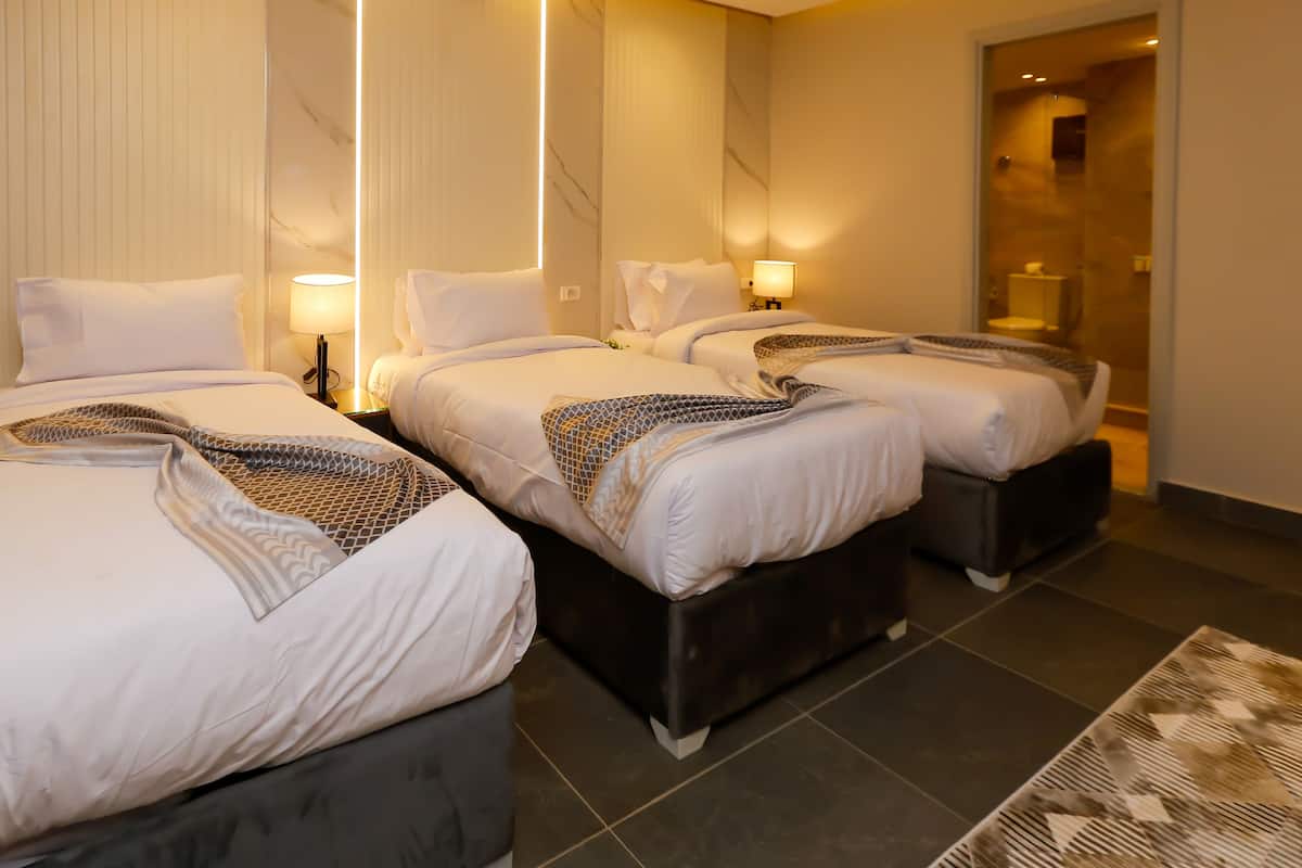 A hotel room with two beds, each with a headboard, a bedside table, and a lamp. The room has a marble wall and a glass shower area.
