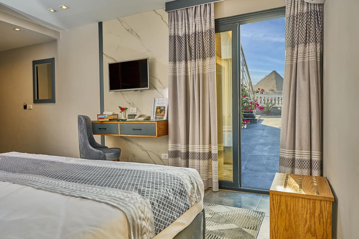 A hotel room with a bed, a desk, a chair, a TV, and a view of the pyramid.