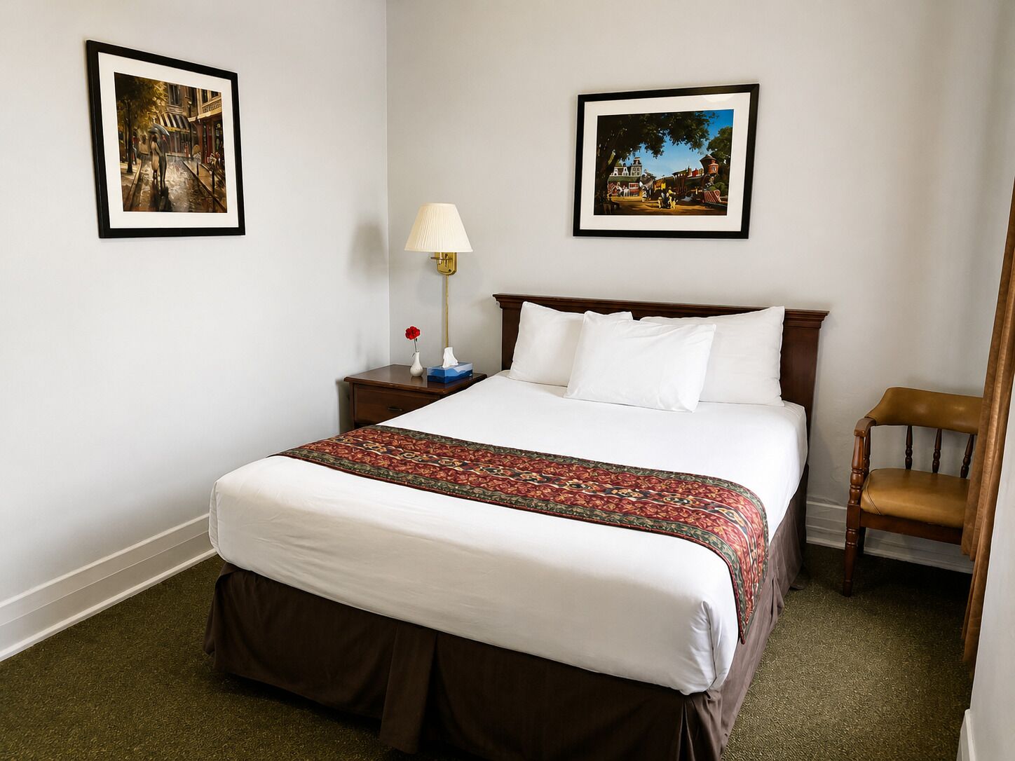 Quarto de hotel com uma cama grande, uma mesinha de cabeceira com abajur, duas imagens emolduradas na parede e uma cadeira de madeira.