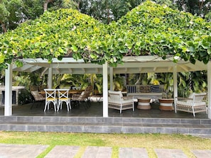 Ein überdachter Außenbereich mit Korbmöbeln und einer Pergola, die von Kletterpflanzen überwuchert ist.