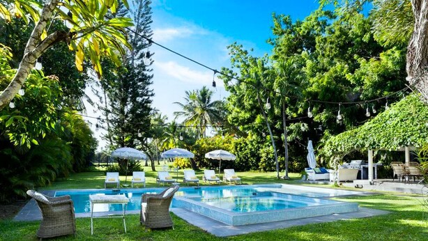 Una piscina rodeada de exuberante vegetación, mobiliario exterior y sombrillas.