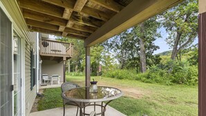 Un patio couvert avec une table et des chaises en verre, une terrasse en bois et une vue sur un jardin luxuriant et des arbres.