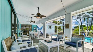 A spacious outdoor lounge area with white furniture, ceiling fans, string lights, and a view of the pool and greenery.