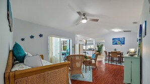 A spacious living area with a wooden floor, a dining table with chairs, a wicker sofa, and a ceiling fan.
