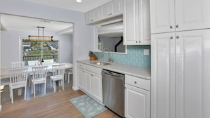 A modern kitchen with white cabinetry, a stainless steel dishwasher, and a window overlooking a garden. - Waterfront Property E. Quogue Minutes From The Hamptons (East Quogue)