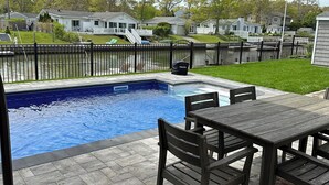 A swimming pool with a patio area, including a table and chairs, and a red umbrella. - Waterfront Property E. Quogue Minutes From The Hamptons (East Quogue)