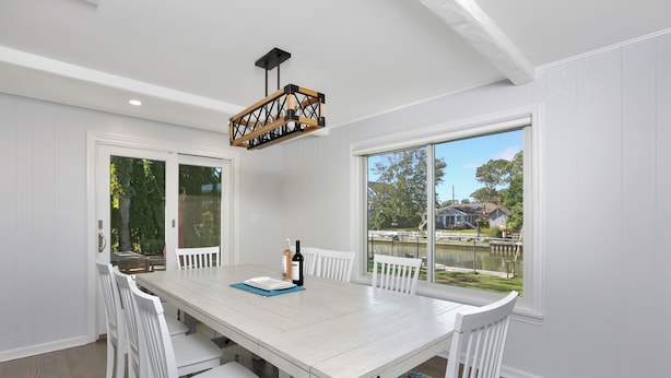 A dining area with a white table and chairs, a wooden pendant light, and a view of a lake and houses through large windows. - Waterfront Property E. Quogue Minutes From The Hamptons (East Quogue)