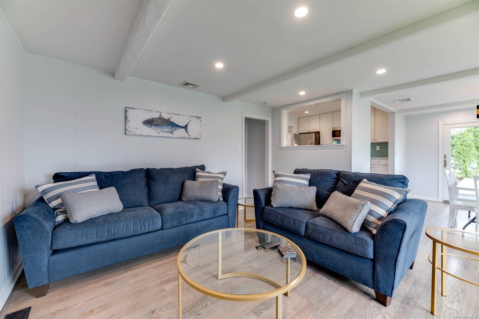 A living room with two blue sofas, a glass coffee table, and a kitchen visible through an open door.