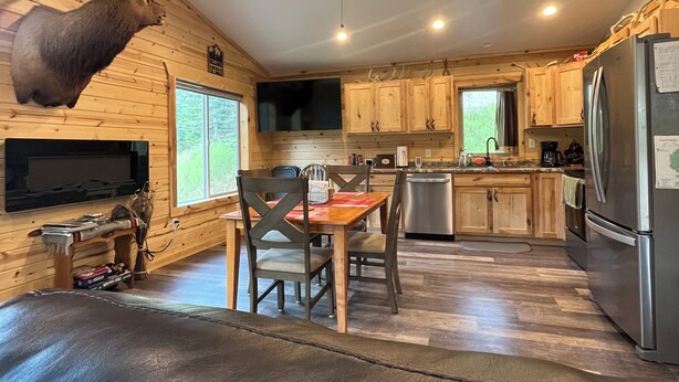 A cabin interior with a dining table, chairs, a ceiling fan, a mounted deer head, wooden cabinets, a refrigerator, and a fireplace. - Beautiful Cabin located in the Little Belt Mountains (White Sulphur Springs)