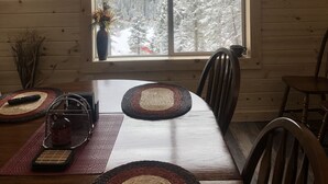 A wooden dining table with chairs, a vase with dried plants, and a view of snow-covered mountains through a large window. - Beautiful Cabin located in the Little Belt Mountains (White Sulphur Springs)