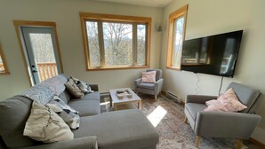 A modern living room with a grey sofa, two armchairs, a coffee table, and a flat-screen TV mounted on the wall. Large windows provide natural light and a view of the outdoors. - Private mountain view haven in the beautiful West Kootenays (Kaslo)