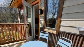 A wooden deck with a round table and two chairs, a wooden railing, and a window with a view of trees. - Private mountain view haven in the beautiful West Kootenays (Kaslo)