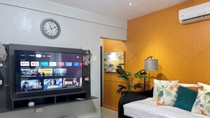 A modern bedroom with a TV mounted on the wall displaying streaming apps, a bed with floral pillows, a grey ottoman, and a wall-mounted clock.