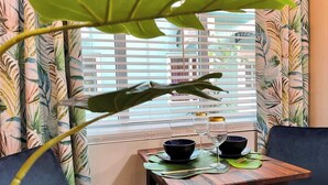 A dining area with a table set for two, featuring black bowls and wine glasses, and a large plant in the foreground.