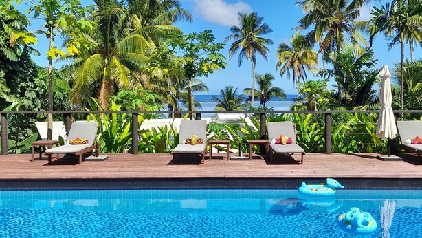 A poolside area with lounge chairs and umbrellas, surrounded by palm trees and a view of the ocean. - Kaila Na Ua Resort (Korotogo)